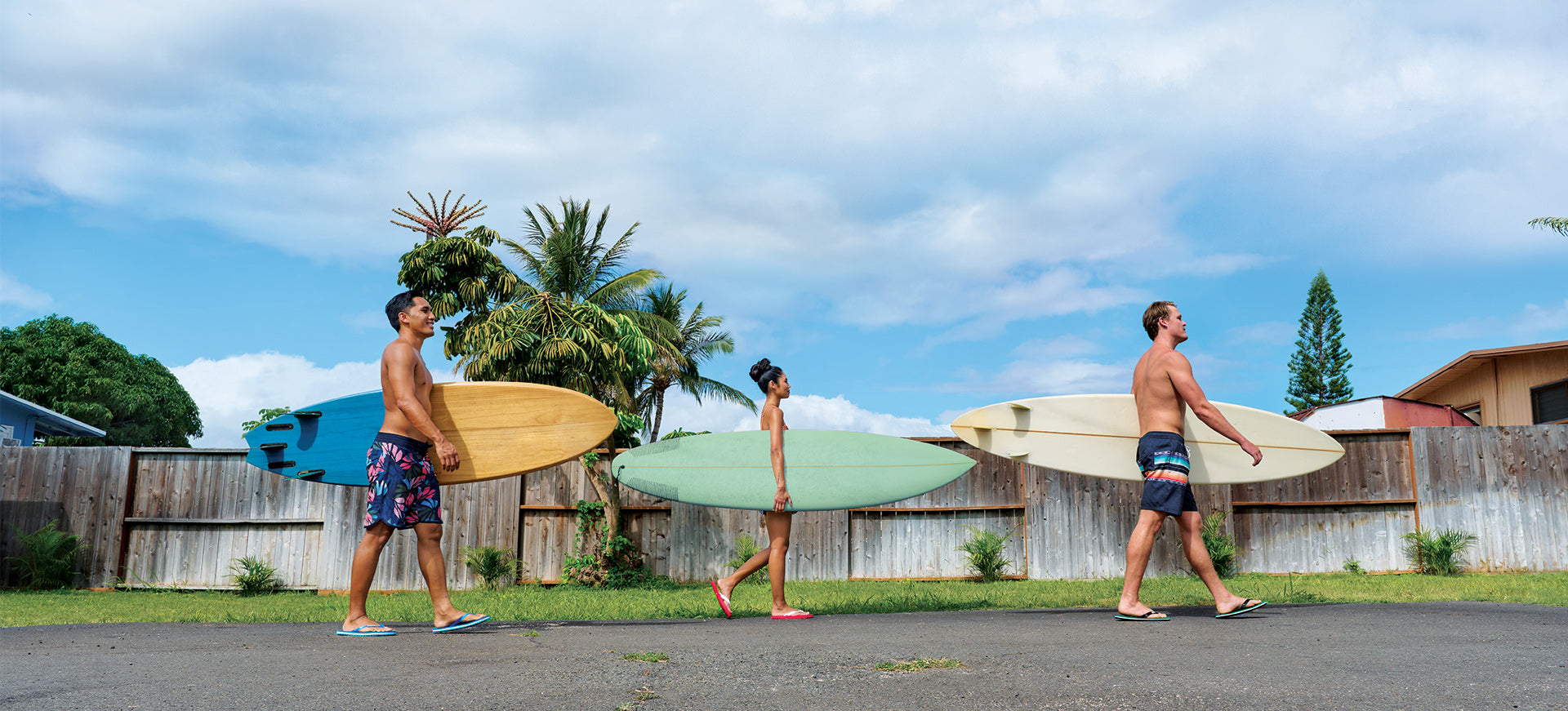 3 surfers carrying boards walking on a street wearing Locals slippahs