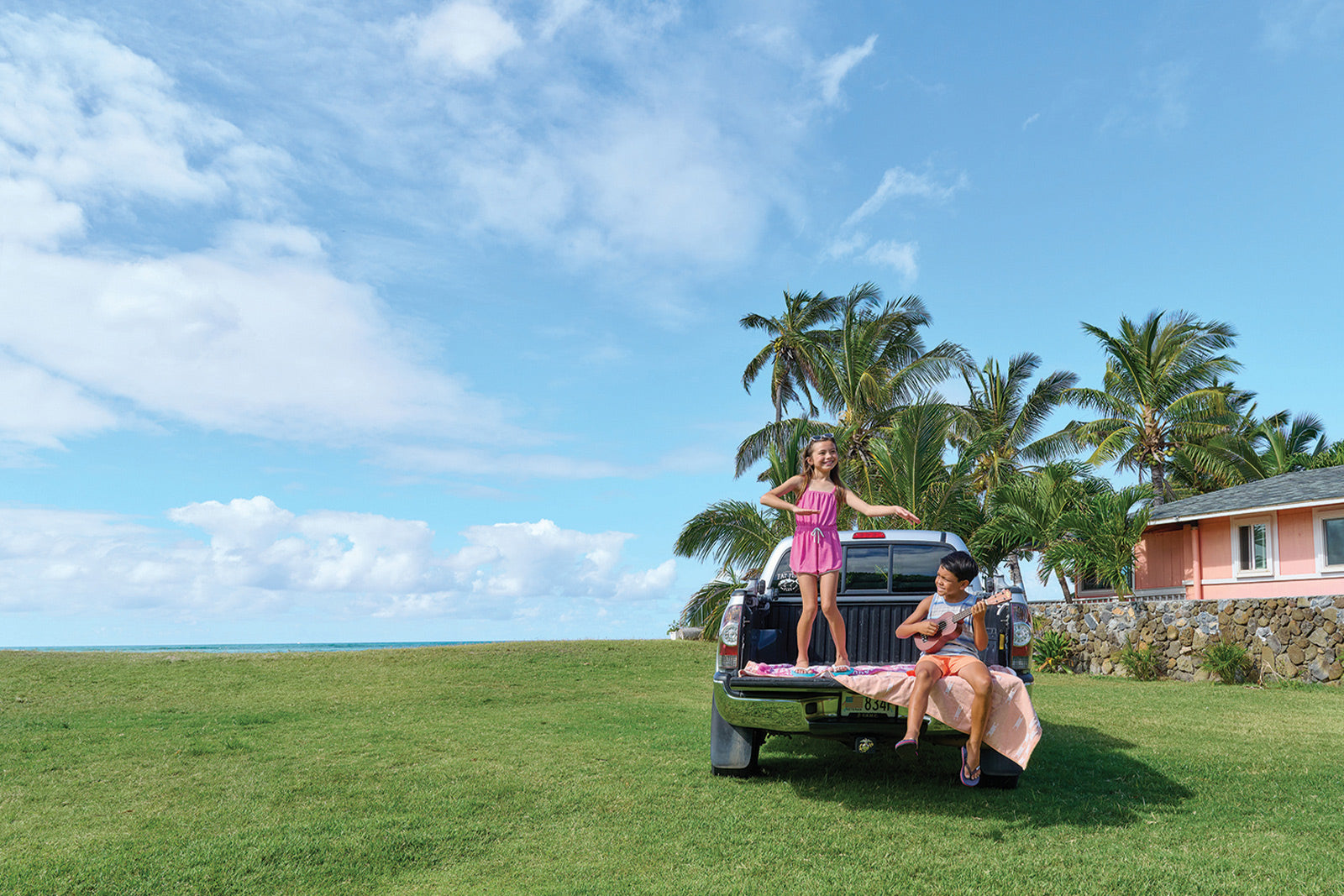 Children playing ukelele and hula in back of truck on lawn wearing Locals slippahs