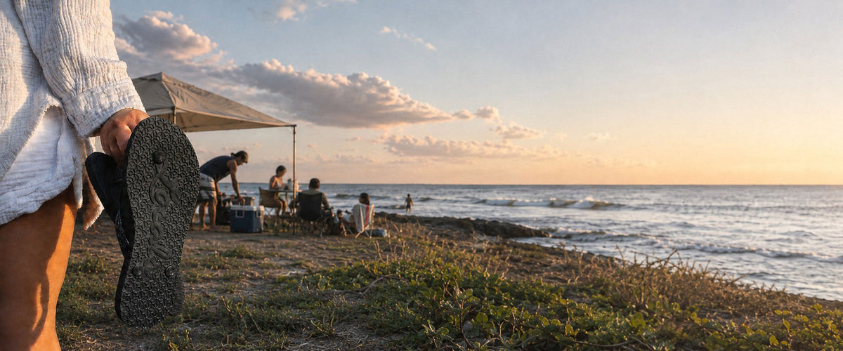 A person in a white ribbed sweater holds up a Locals flip-flopsl on a beach shoreline at sunset, with ocean waves, a small beach setup under a canopy, and seated people in the background.