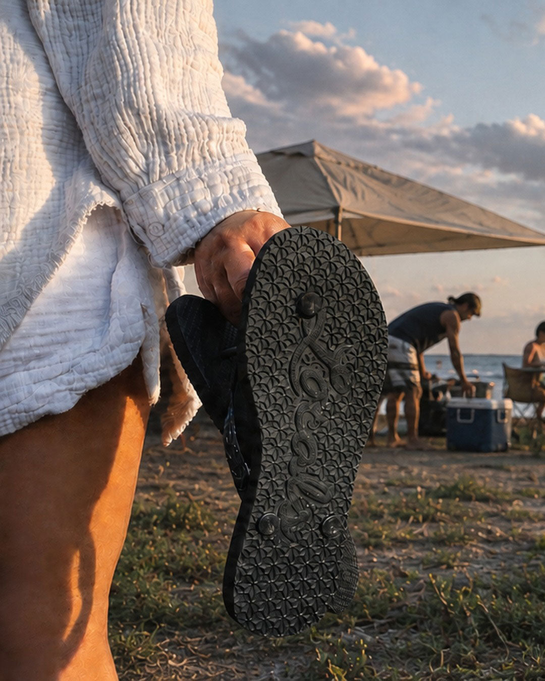 A person in a white ribbed sweater holds up a Locals flip-flopsl on a beach shoreline at sunset, with ocean waves, a small beach setup under a canopy, and seated people in the background.