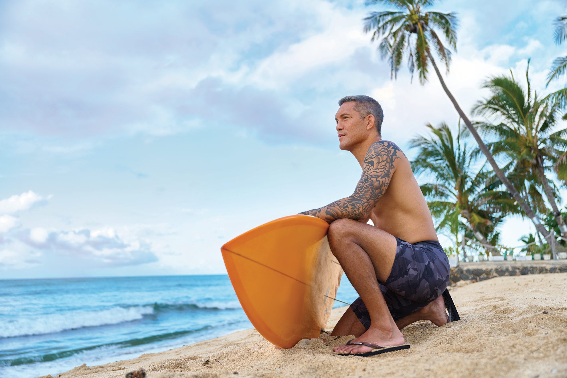 Man sitting on a beach holding a yellow surfboard wearing Locals slippahs