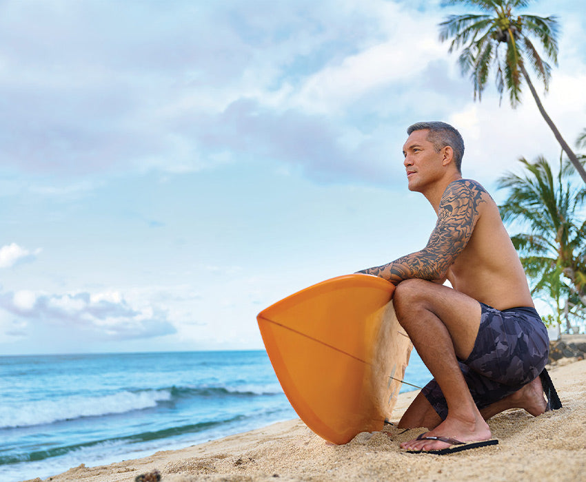 Man sitting on a beach holding a yellow surfboard wearing Locals slippahs
